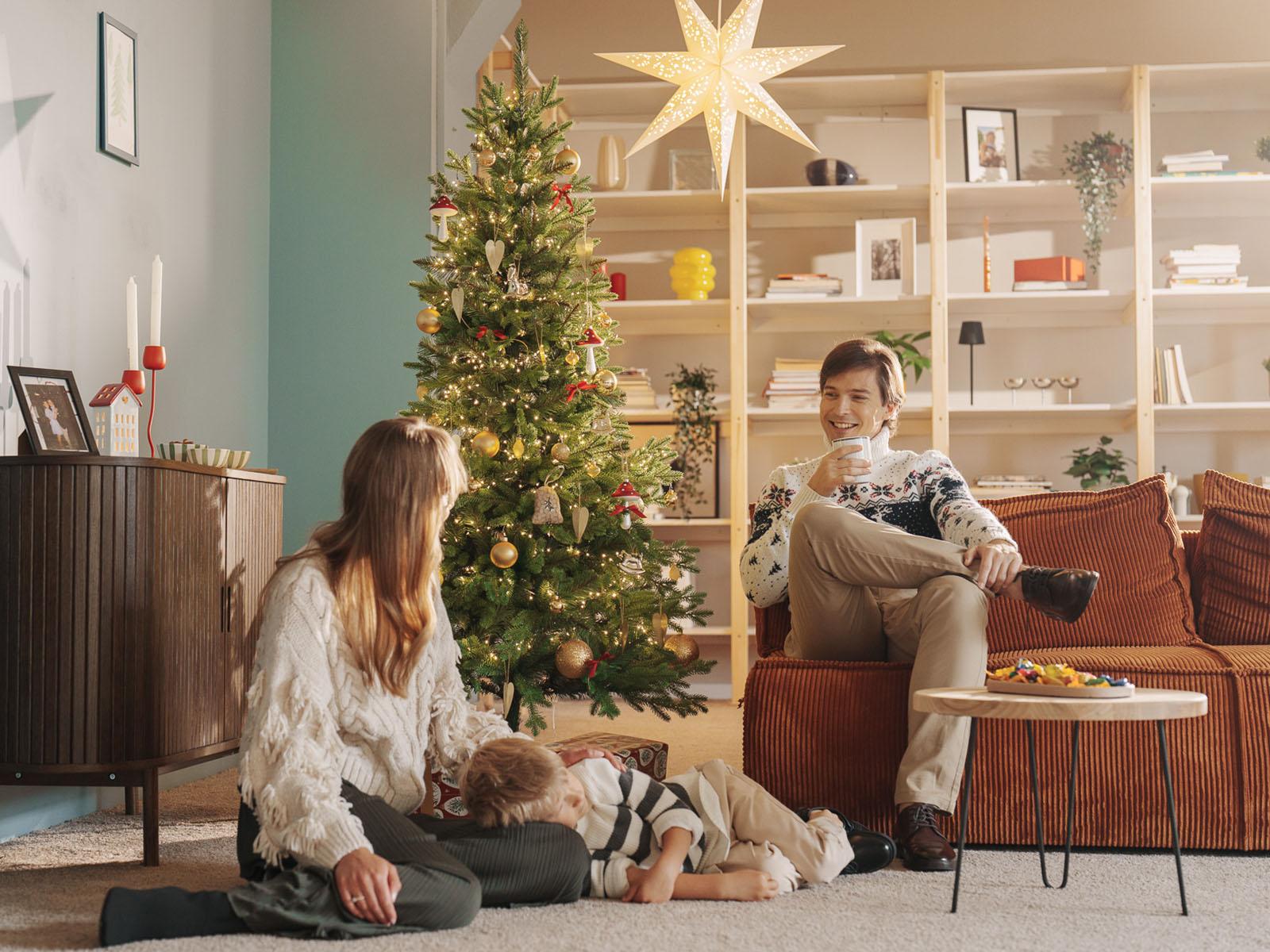 Tata siedzi na sofie z kubkiem kawy w ręku, mama siedzi na podłodze, synek śpi z głową na jej kolanach, a w tle stoi sztuczna jodła kaukaska slim FairyTrees FT05, tworząc spokojny, cichy nastrój.