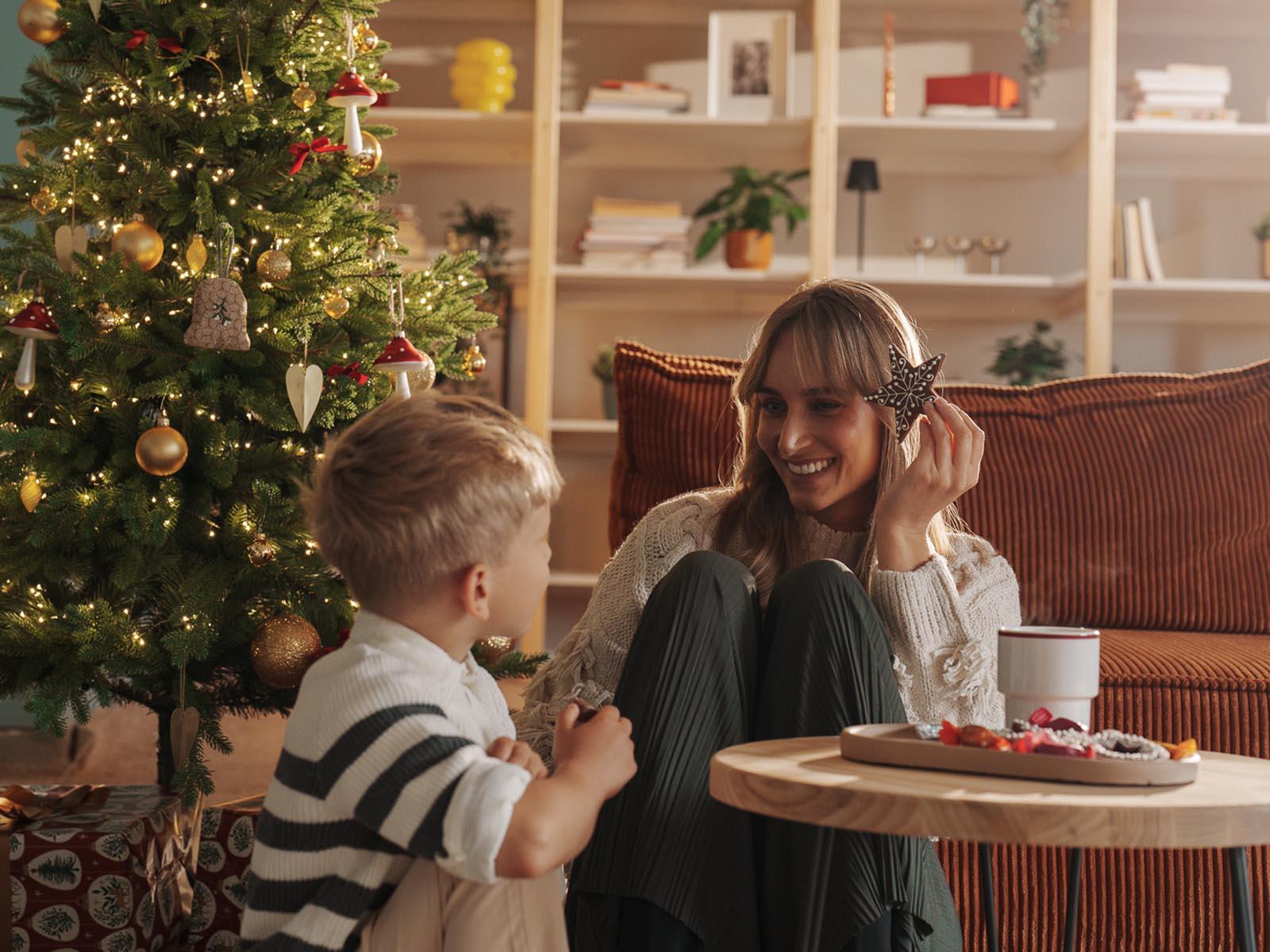 Mama siedzi z synem na podłodze przy udekorowanej sztucznej choince FairyTrees FT05 i prezentach w przytulnym salonie.