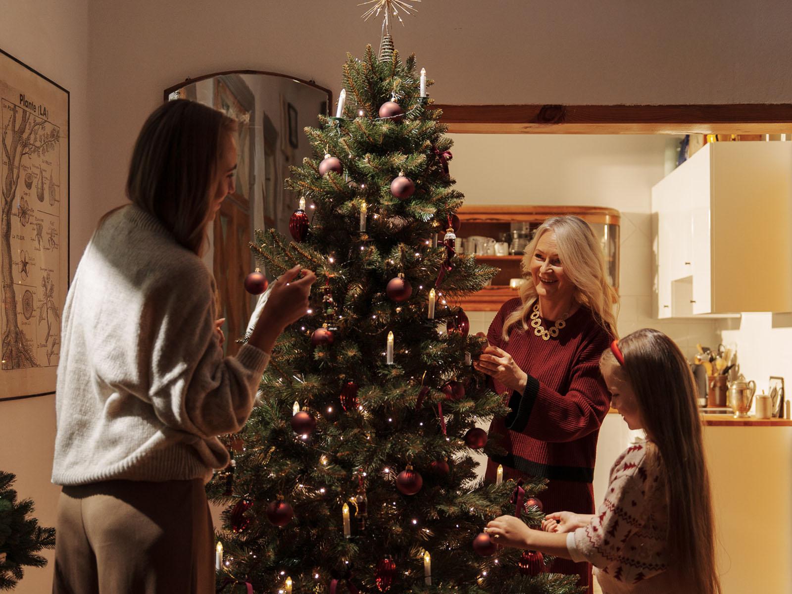 Babcia, mama i wnuczka wspólnie ubierają sztuczną jodłę kaukaską FairyTrees FT14 w salonie, a w tle widać otwartą kuchnię.